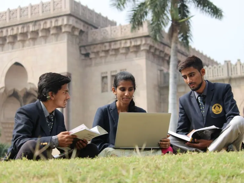 Indian male and female students studying together on a lawn in front of a grand university building
